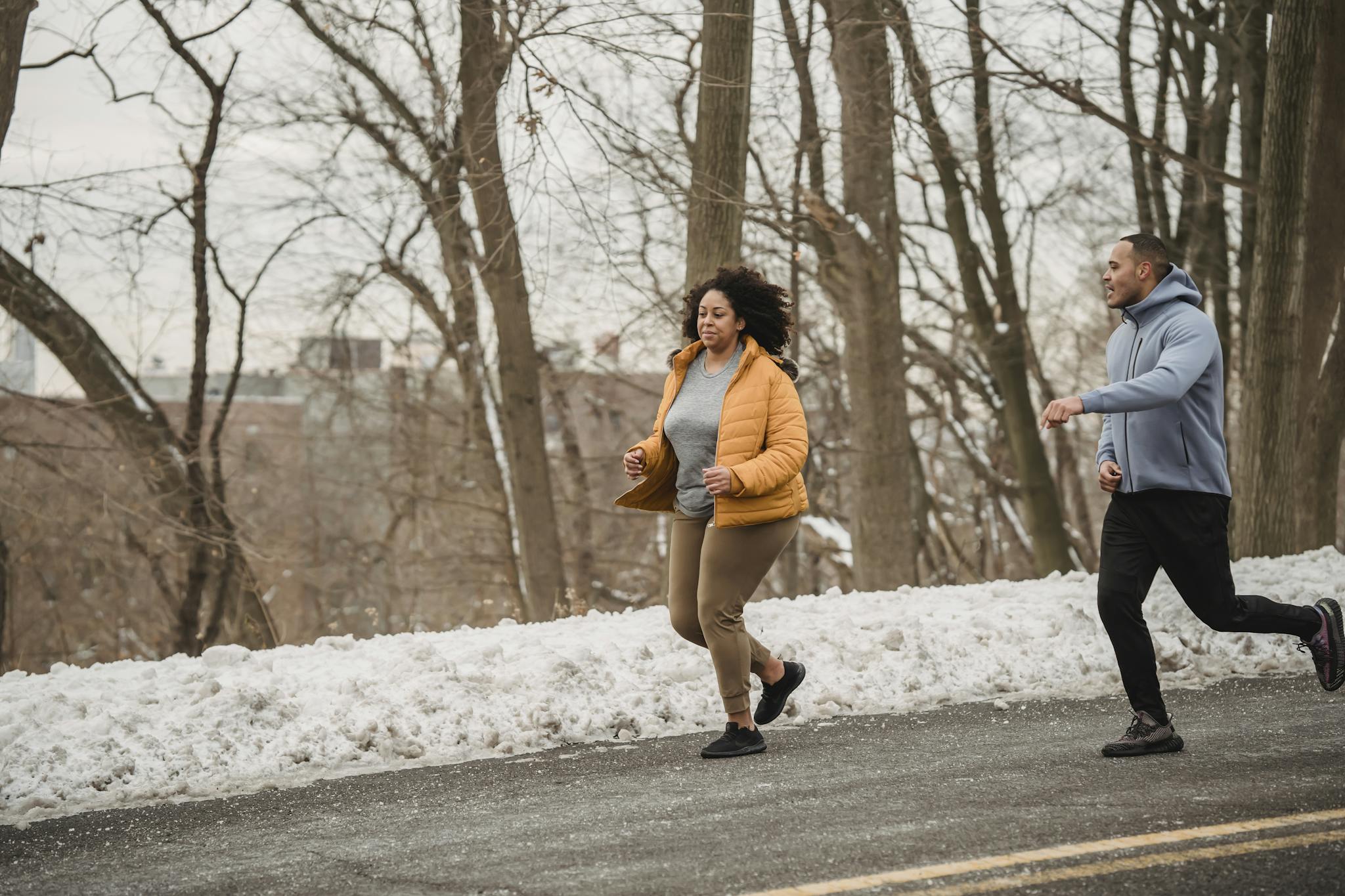 Full body of plus size African American female running with personal instructor on asphalt road during winter training on street